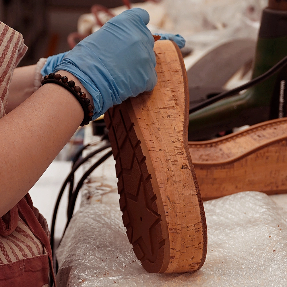 Una persona che indossa un guanto e un braccialetto lavora su una suola di sughero, circondata da strumenti e materiali su un banco di lavoro ricoperto di pluriball.