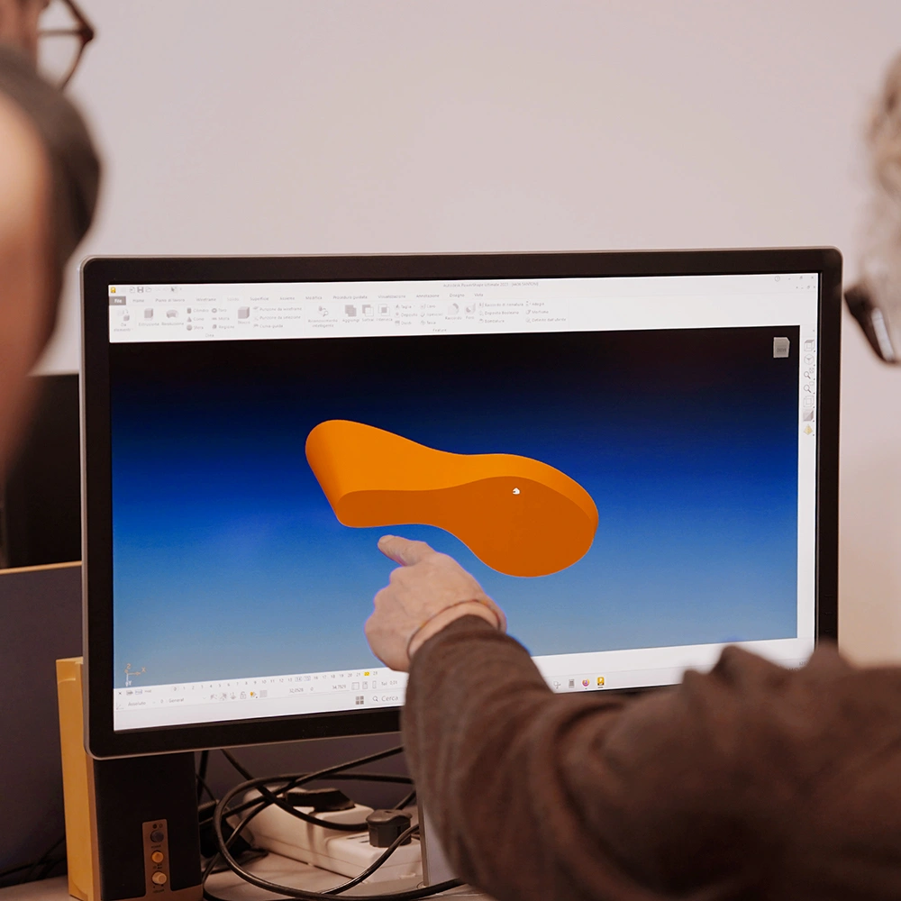 Due persone guardano il monitor di un computer che visualizza un programma di modellazione 3D con un oggetto arancione, forse una suola di scarpa, su uno sfondo blu. Una persona indica lo schermo.
