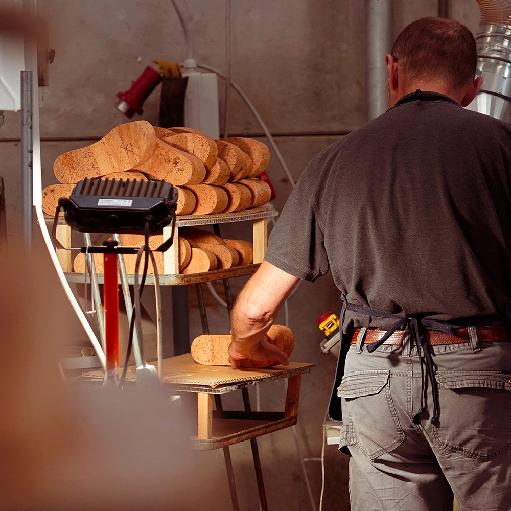 Una persona in un laboratorio seleziona grandi pezzi di sughero da uno scaffale, indossando una camicia scura e un grembiule. Gli scaffali contengono diversi pezzi di sughero e sullo sfondo sono visibili gli strumenti del laboratorio.
