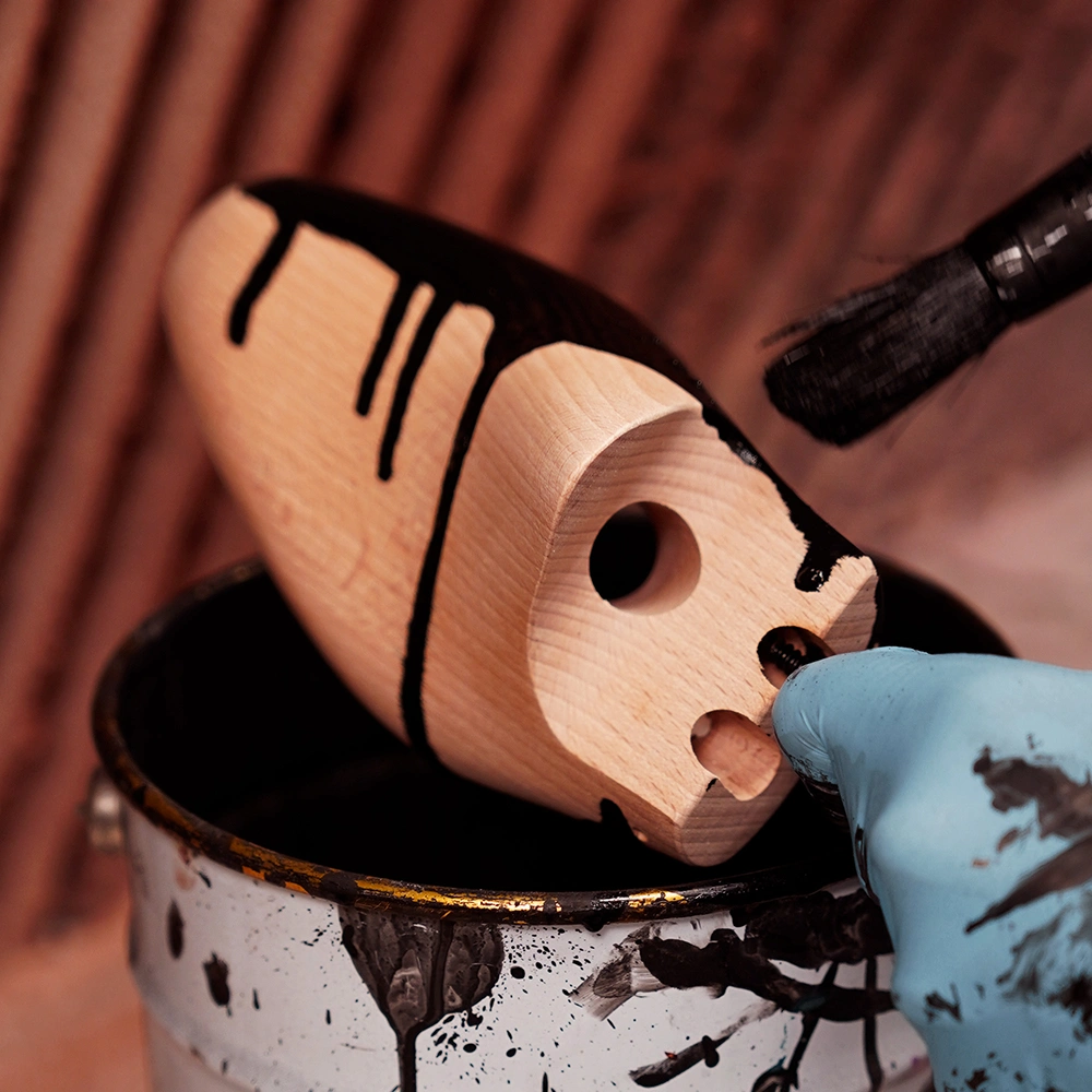 Una persona con un guanto blu dipinge di nero un prototipo in legno, tenendolo sopra un barattolo di vernice con la vernice nera che cola dal pennello e dal prototipo.