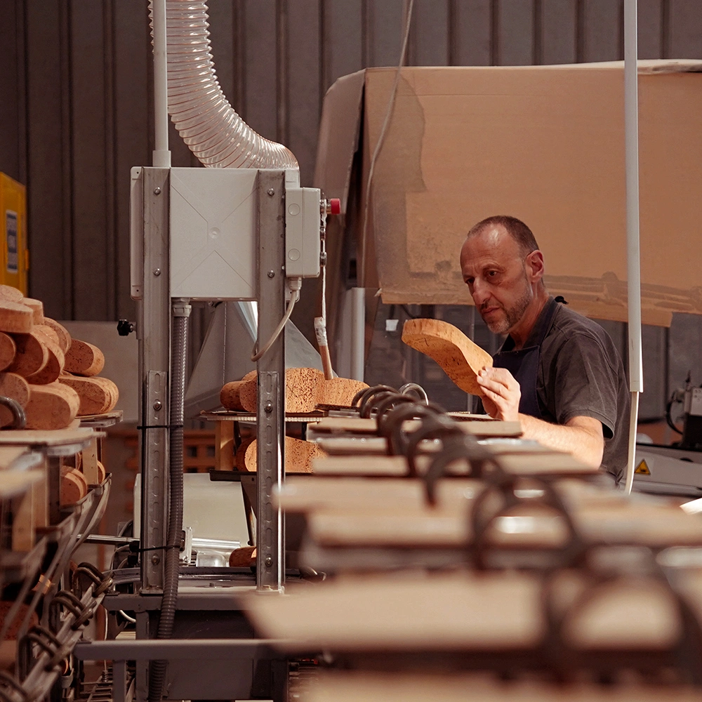 Un uomo in un'officina ispeziona una suola di sandalo in sughero, circondato da macchinari e file di suole in sughero, in un ambiente di fabbrica con luci soffuse.