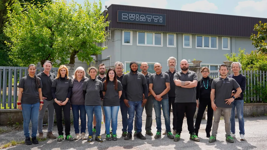 Un gruppo di quindici persone è in fila all'aperto davanti a un edificio industriale grigio con l'insegna Buiatti. Tutti indossano camicie grigie e scarpe verdi, sono rivolti verso la telecamera e sorridono. Sullo sfondo ci sono alberi e una recinzione.
