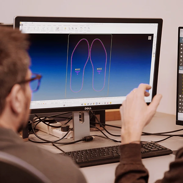 Due persone sedute a una scrivania guardano lo schermo di un computer che mostra un progetto CAD di due solette per scarpe. Una persona fa un gesto verso lo schermo, discutendo del progetto. Sulla scrivania sono visibili un altro monitor e degli accessori per computer.